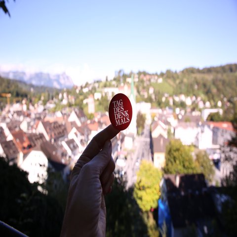 Auf einem Finger klebt ein runder Sticker mit der Aufschrift "Tag des Denkmals", im Hintergrund ist eine Stadt zu sehen