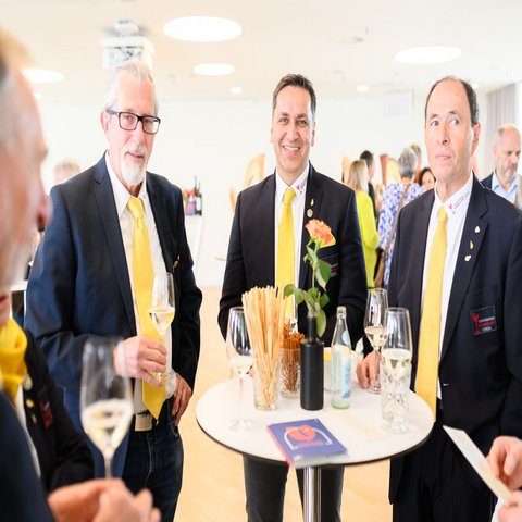 Drei Männer mit Anzug und gelber Krawatte stehen an einem Stehtisch