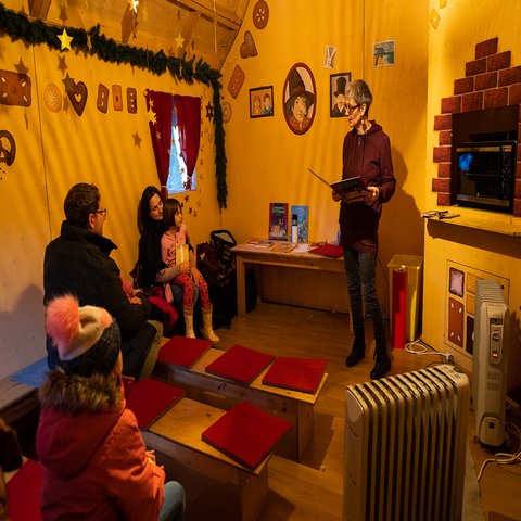 Eine Frau liest aus einem Buch vor, zwei Eltern und ihre Kinder lauschen